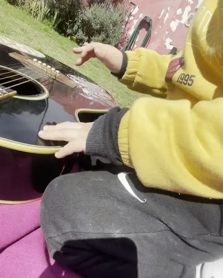 Las manos de un niño con buzo amarillo tocando las cuerdas de una guitarra negra sobre una manta violeta.