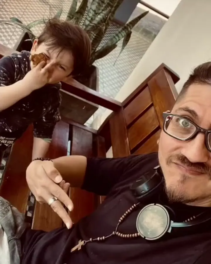Jere con lentes y auriculares al cuello, sonriendo, y su hijo detrás haciendo un gesto con la mano.