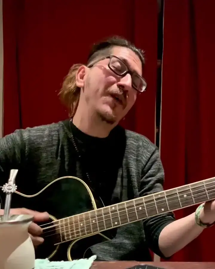 Jere Masih canta y toca la guitarra sentado frente a una cortina roja, con un mate sobre la mesa; se ríe mientras canta.
